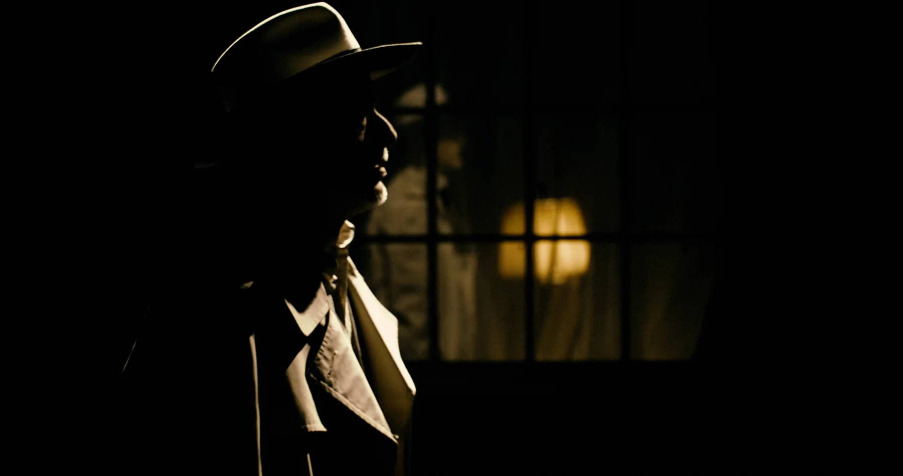 A man in a hat stands in deep shadow beside a window.