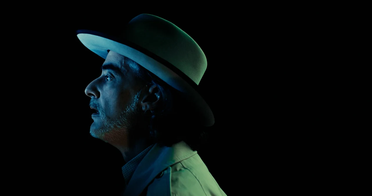 P.T. Mitchell, wearing a light hat, illuminated in cyan against the black void.